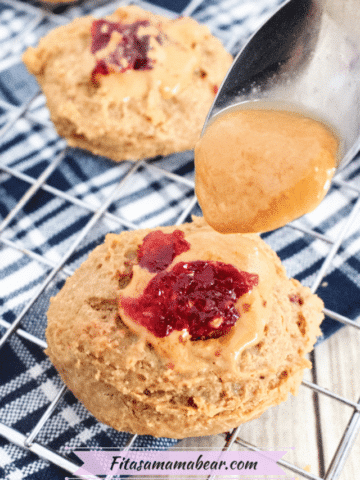 Cooling rack on a blue linen with multiple peanut butter thumbprint cookies topped with jam and a spoon of peanut butter above the first