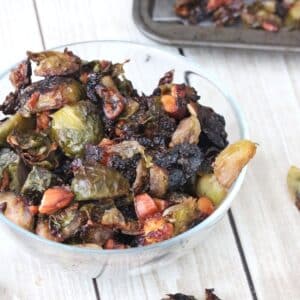 Glass bowl with roasted brussels sprouts and more sprouts on a baking tray behind it.