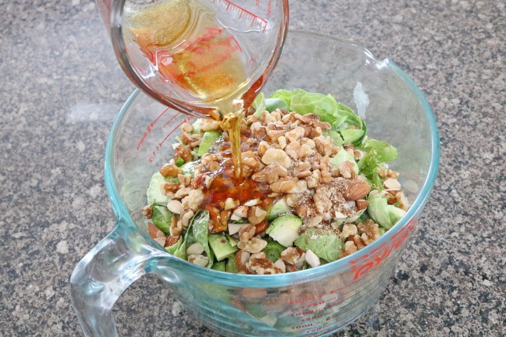 Maple syrup being poured over chopped brussel sprouts and nuts in a measuring cup