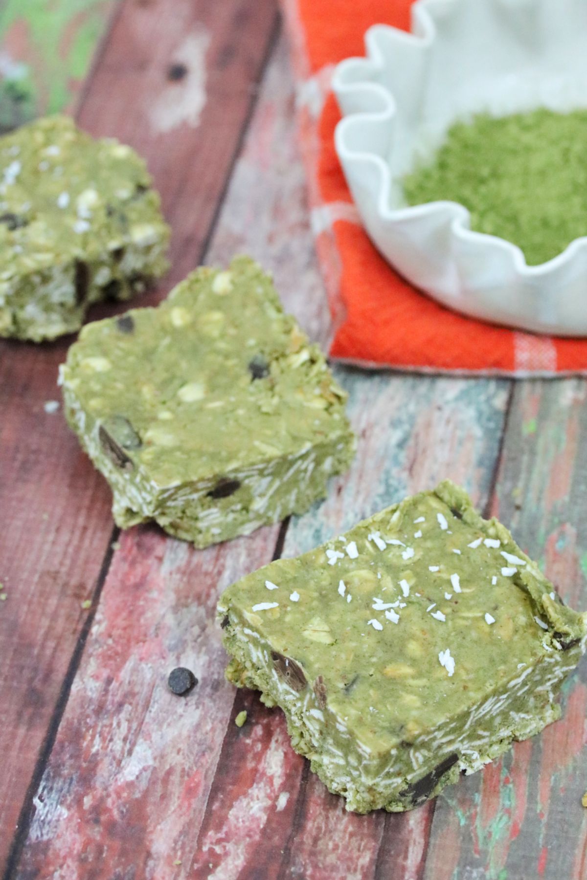 Three green protein bars lined up with a bowl of matchinga powder behind it.