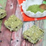 Three green protein bars lined up with a bowl of matchinga powder behind it.