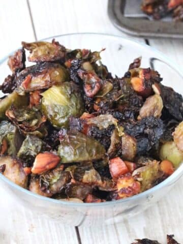 Glass bowl with roasted brussels sprouts and more sprouts on a baking tray behind it.