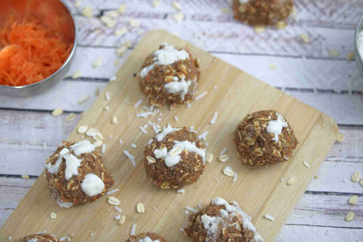 Carrot cake protein balls on a wooden cutting board with a bowl of carrot beside them and coconut and oats around them.