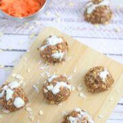 Carrot cake protein balls on a wooden cutting board with a bowl of carrot beside them and coconut and oats around them.