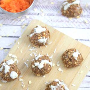 Carrot cake protein balls on a wooden cutting board with a bowl of carrot beside them and coconut and oats around them.