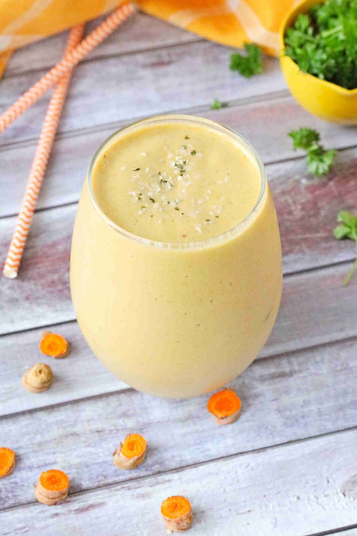 Mango carrot turmeric smoothie in a clear glass with a yellow bowl of parsley, orange straws and sliced turmeric around/behind it.