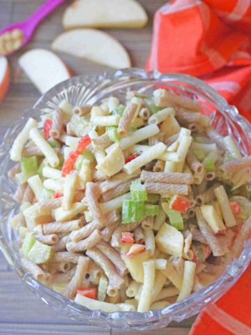 Glass bowl with macaroni salad in it with celery and red peppers and apple slices and an orange linen behind it.