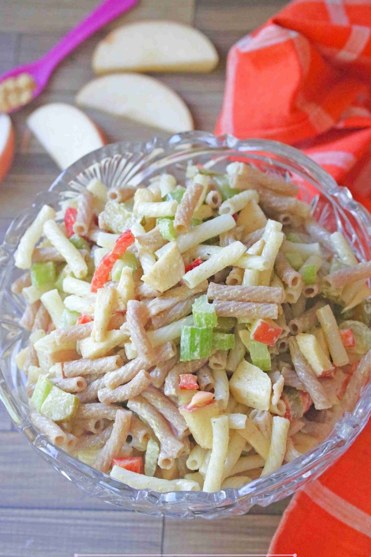 Glass bowl with macaroni salad in it with celery and red peppers and apple slices and an orange linen behind it.