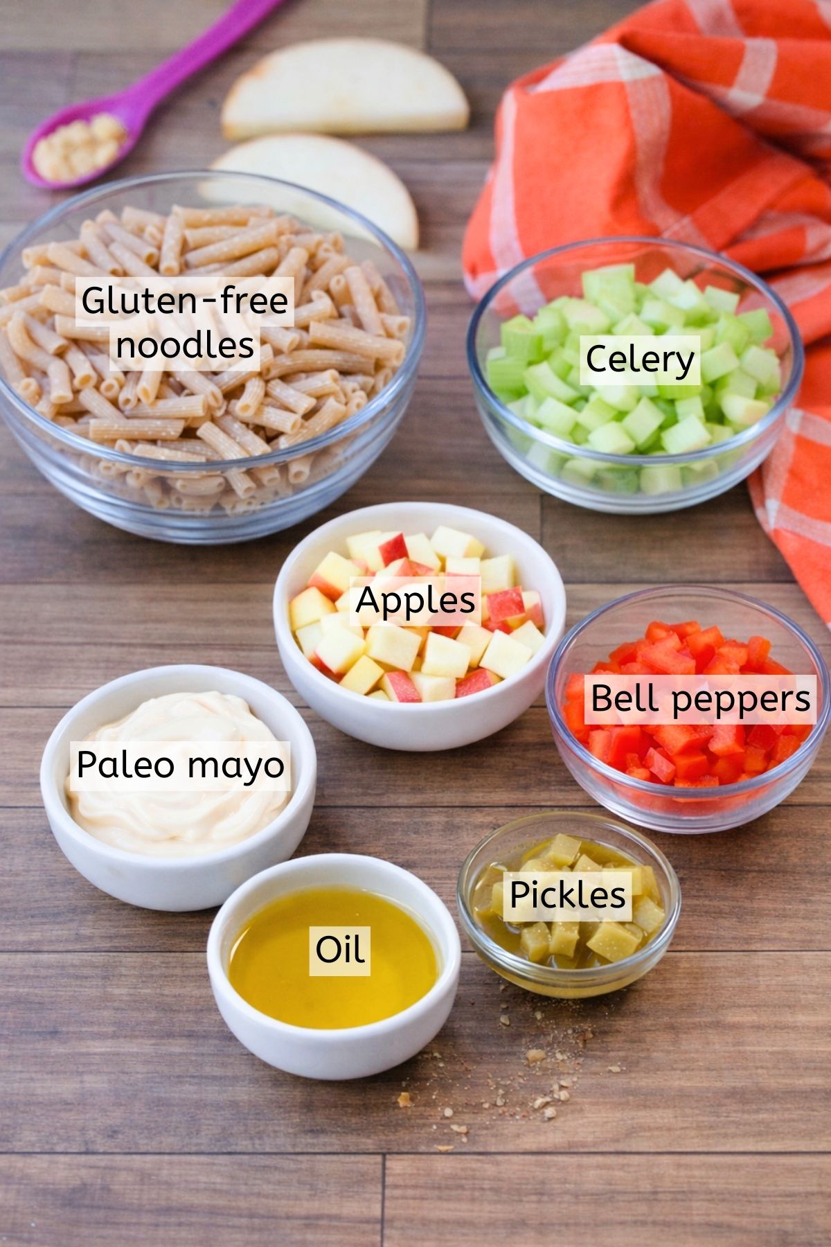 Apples, celery, mayonnaise and noodles in glass bowls with text labels over top.