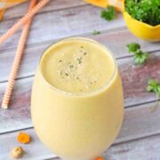 Gourmet mango carrot turmeric smoothie in a clear glass with a yellow bowl of parsley, orange straws and sliced turmeric around/behind it
