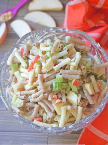 Glass bowl with macaroni salad in it with celery and red peppers and apple slices and an orange linen behind it