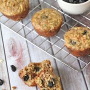 One morning glory muffin split in half with more muffins and a bowl of dried fruit on a cooling rack behind it.
