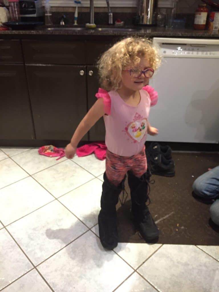 Toddler in a pink dress, orange pants and her moms boots standing in the kitchen