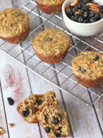 One morning glory muffin split in half with more muffins and a bowl of dried fruit on a cooling rack behind it