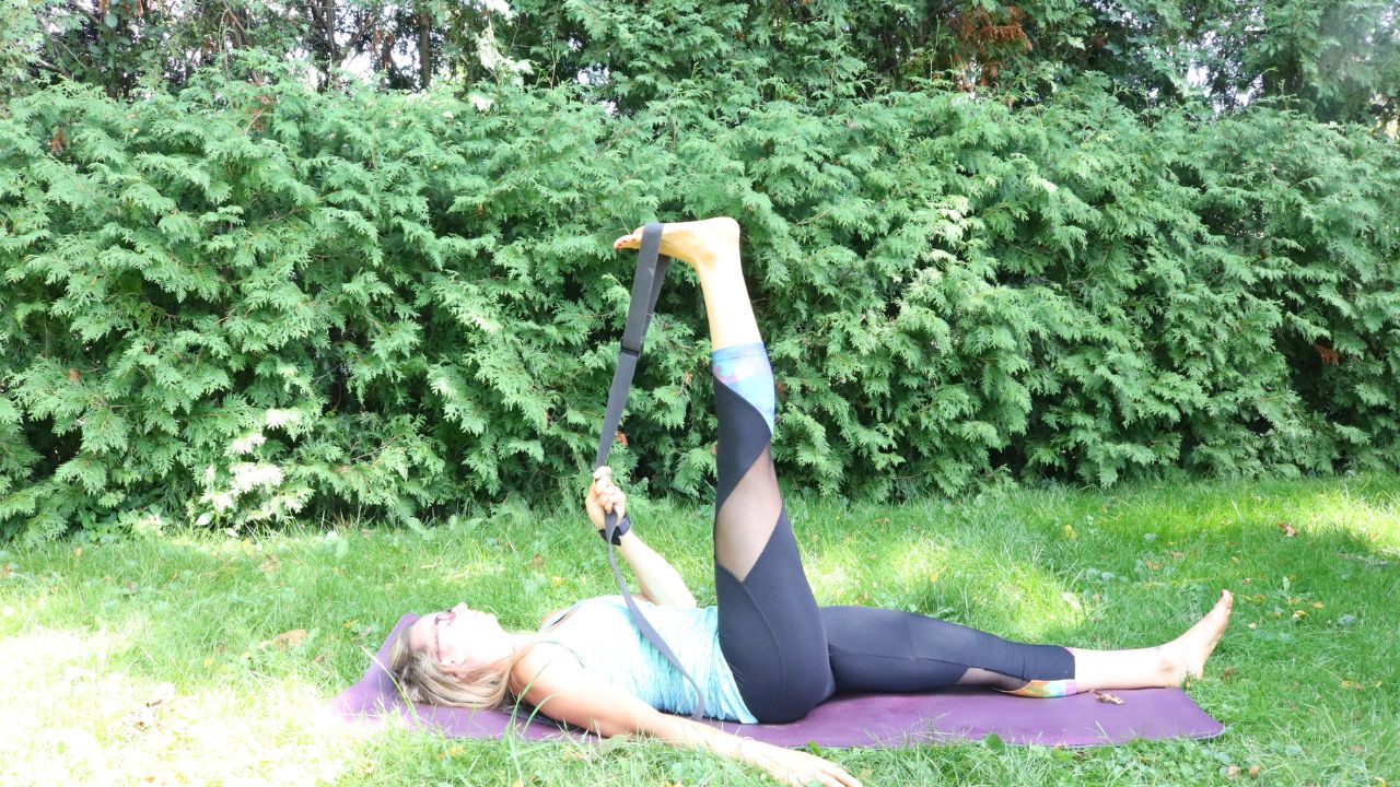 Women laying on back on yoga mat outside stretching one leg with a yoga strap