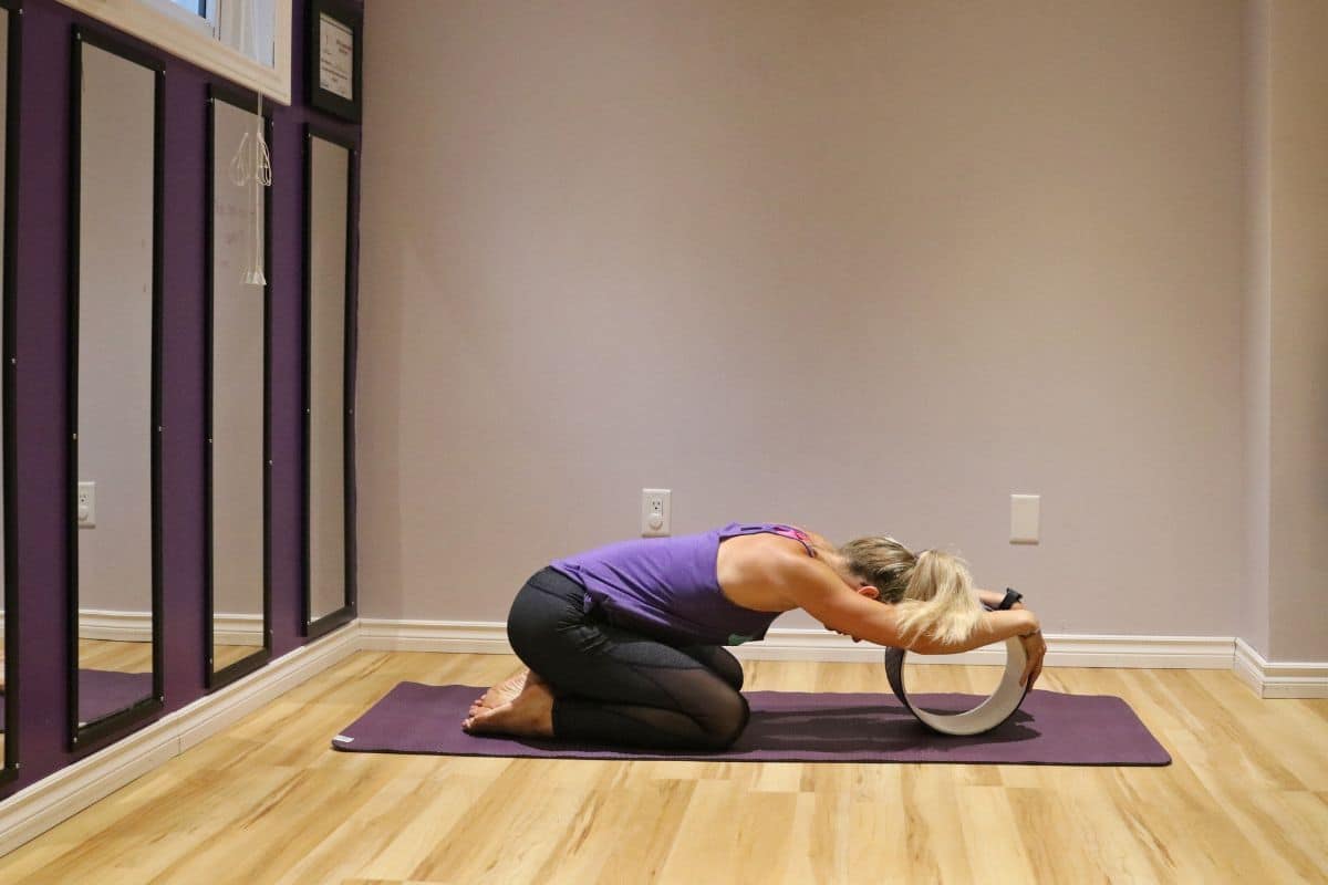 A woman in a purple shirt and black pants performing child's pose on a yoga mat inside with a yoga wheel
