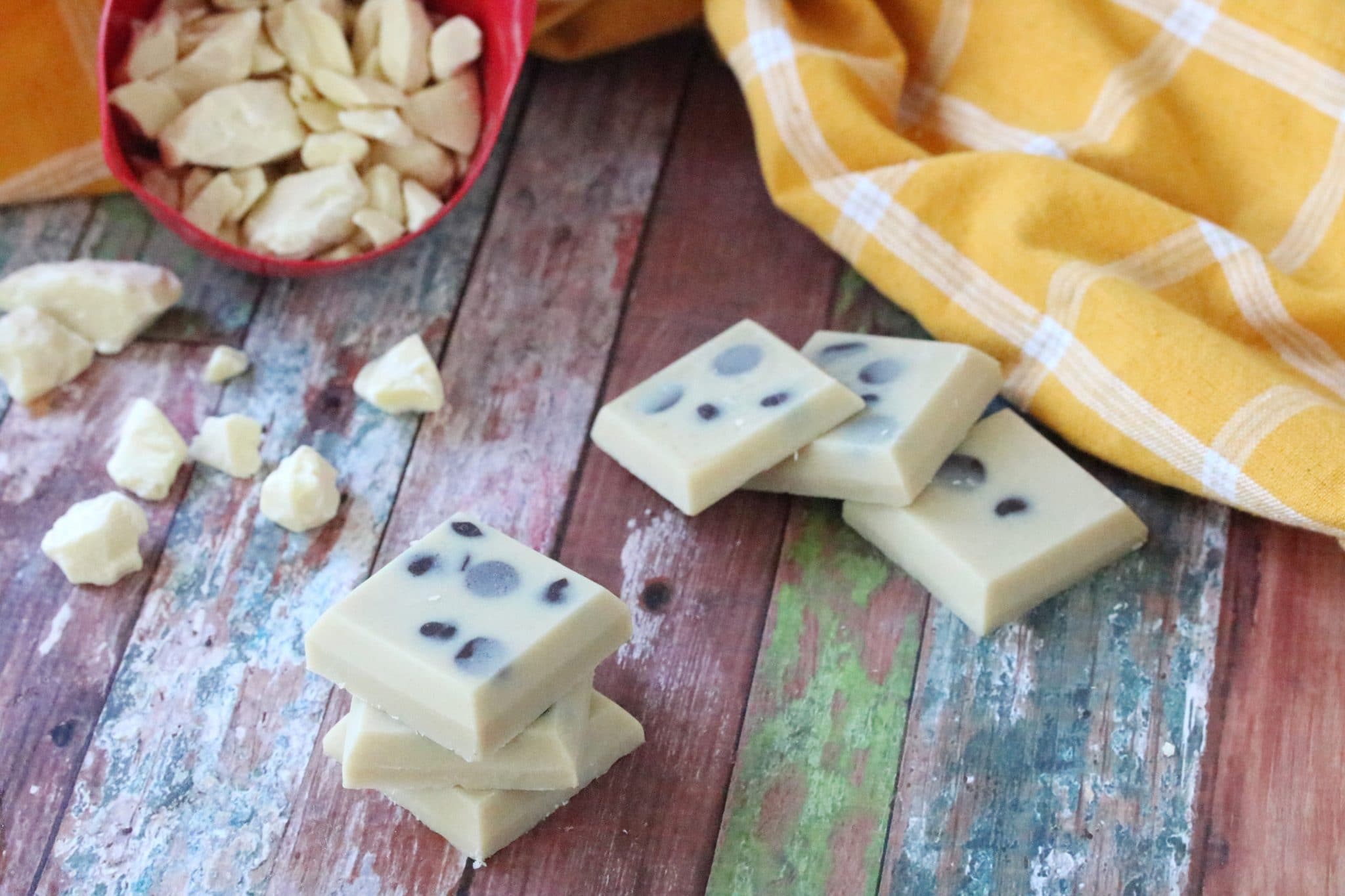 White chocolatye fudge, three squares stacked with three sqaures on table behind it and cacao butter falling out of a red measuring cup