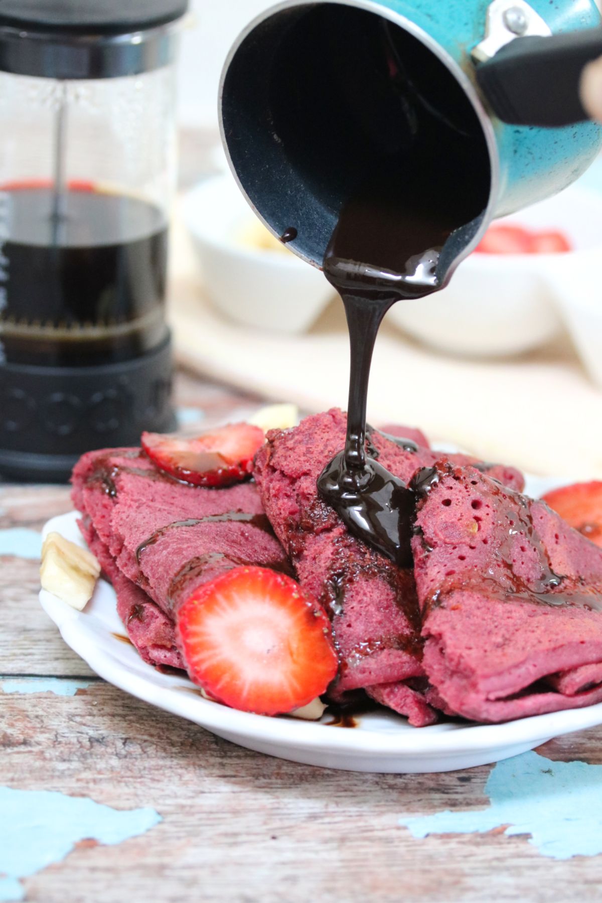 Three pink eggless crepes on a white plate with chocolate sauce being poured on top