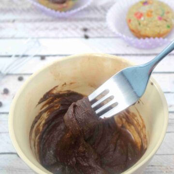 Fork in a bowl of chocolate dairy-free frosting and cupcakes behind the bowl.