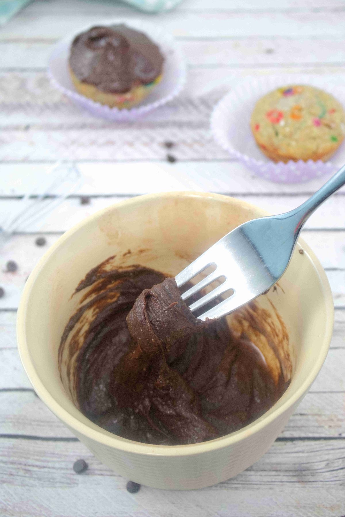 Fork in a bowl of chocolate dairy-free frosting and cupcakes behind the bowl.