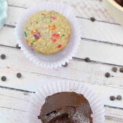 One chocolate frosted cupcake in a purple muffin liner with another cupcake, bowl of chocolate frosting and blue linen behind it.