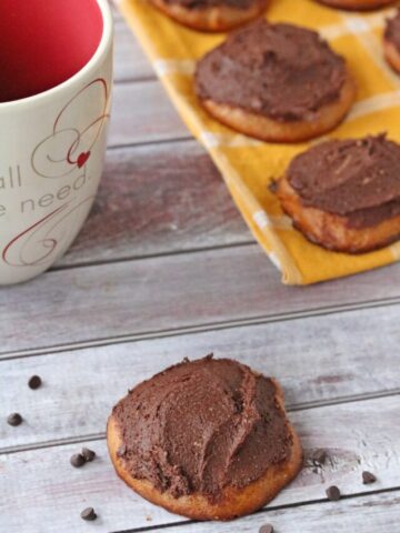 One gluten-free cookie frosted with chocolate surrounded by chocolate chips with a cup of coffee and more cookies on a yellow linen behind it