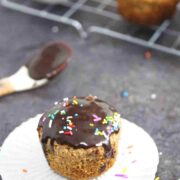 Close up of a vanilla almond flour cupcake with chocolate ganache on top and sprinkles on a white muffin liner with chocolate on a spoon and more cupcakes behind it.