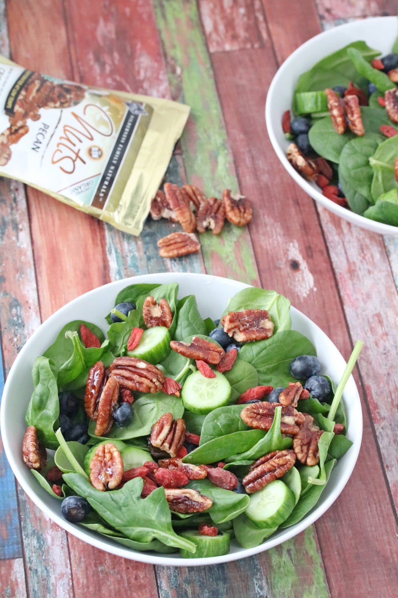 Spinach salad with pecans in a white bowl with another bowl of salad behind it and pecans on the table