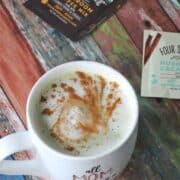 Homemade matcha latte in white mug with cinnamon and matcha and coffee packs around it.