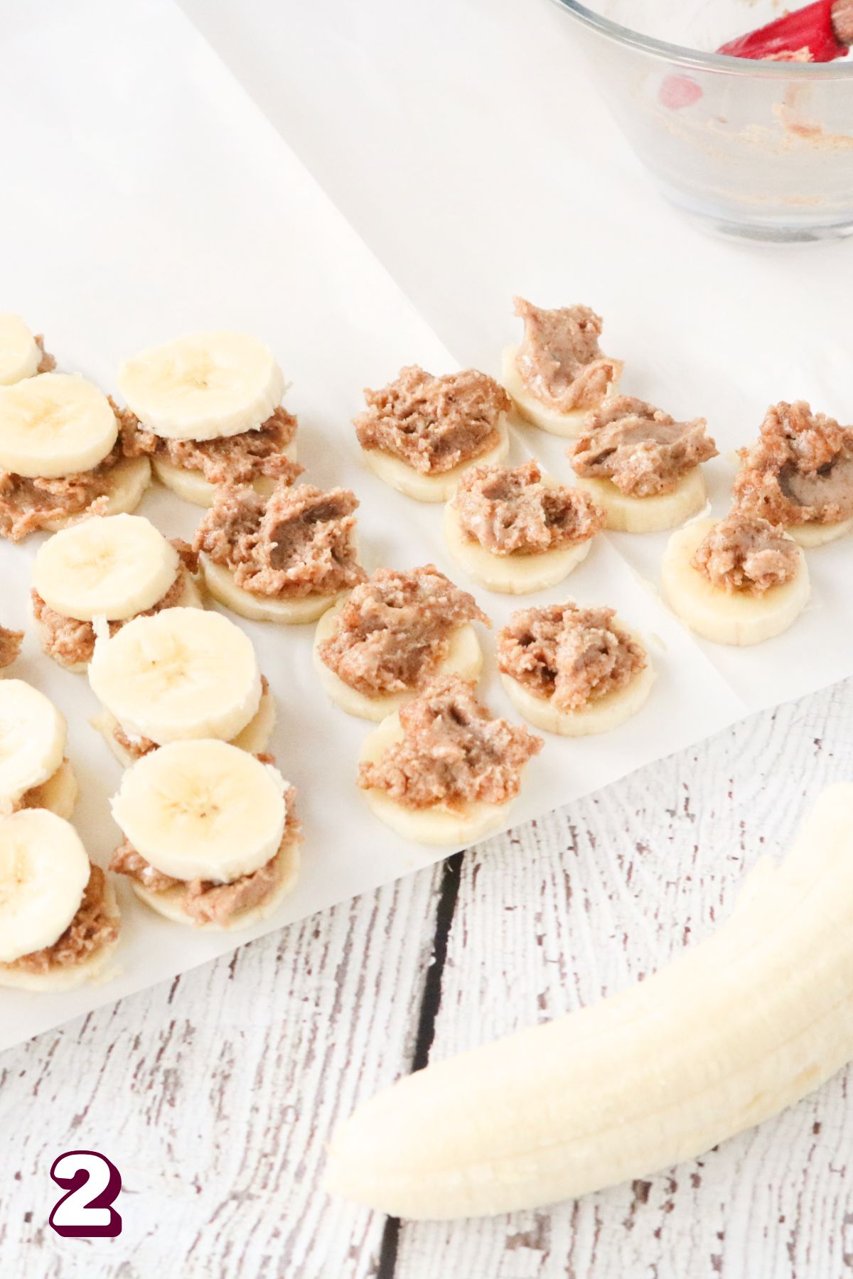 In process image for frozen bananas: sliced bananas on parchment paper with almond butter on top of bananas.