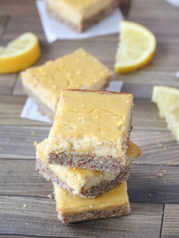 Lemon squares stacked with lemons around them. Lemons in a bowl behind them and more squares around and behind.
