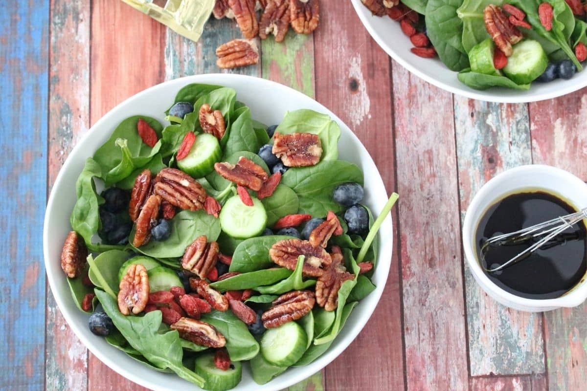 Spinach salad with pecans in a white bowl with another bowl of salad behind it and pecans on the table.