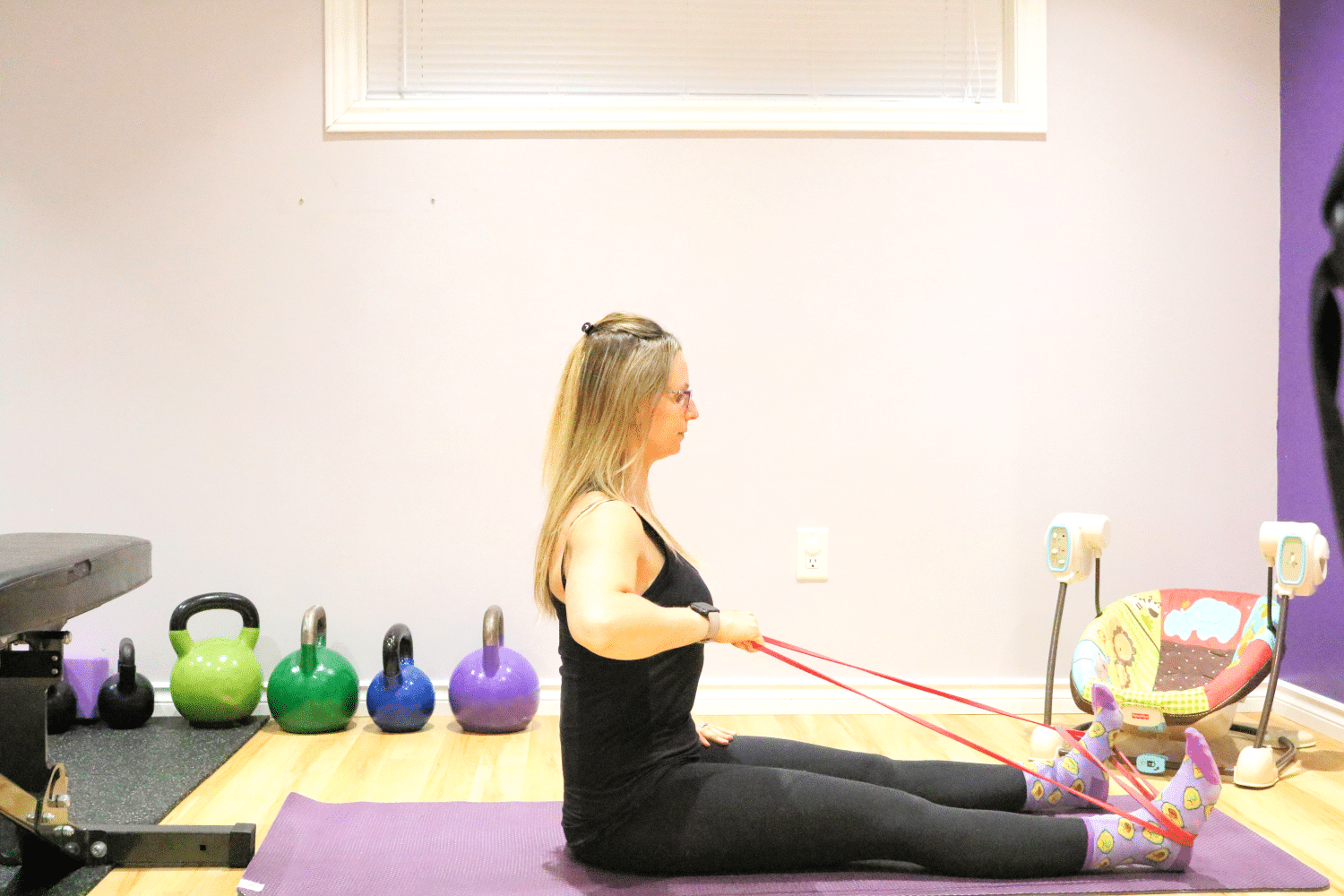 Woman in dark workout clothes sitting on a purple yoga mat performing a seated row with a red resistance band