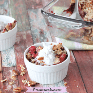 Featured image with text: paleo berry crumble in a white ramekin with pecans around it and more crumble behind