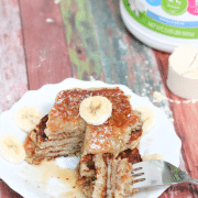 White plate with banana protein pancakes stacked and a slice cut out and on the fork beside the place, banana on and maple syrup top of the pancakes and protein powder behind them