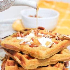 maple syrup being poured onto stacked pumpkin waffles with coconut cream and pecans