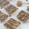 Homemade puffed rice bars topped with chocolate and caramel cut into squares and on parchment paper with a honey stick of almond butter behind them