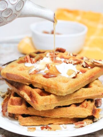 maple syrup being poured onto stacked pumpkin waffles with coconut cream and pecans