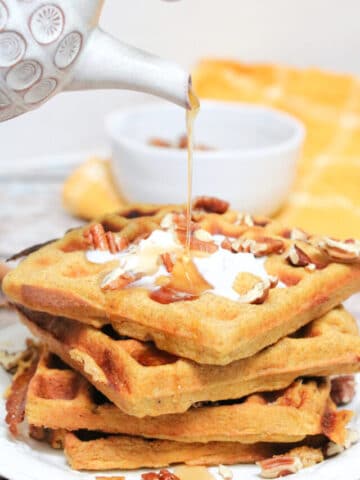 maple syrup being poured onto stacked pumpkin waffles with coconut cream and pecans