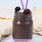 Featured image with text: chocolate cherry smoothie in a mason jar topped with coconut whip, cherries and purple straws, peach napkin behind it