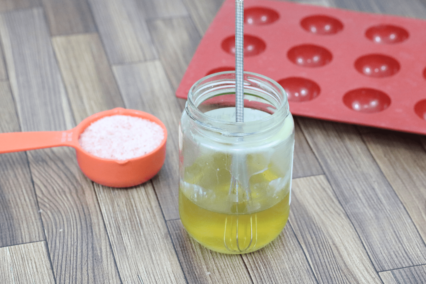 Melted oils and butters in a glass mason jar with a small whisk in it and a cup of pink salt and red silicone mold beghind it