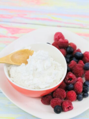 Pink bowl with whole30 whipped cream and a wooden spoon in it and berries on the side of a white plate.