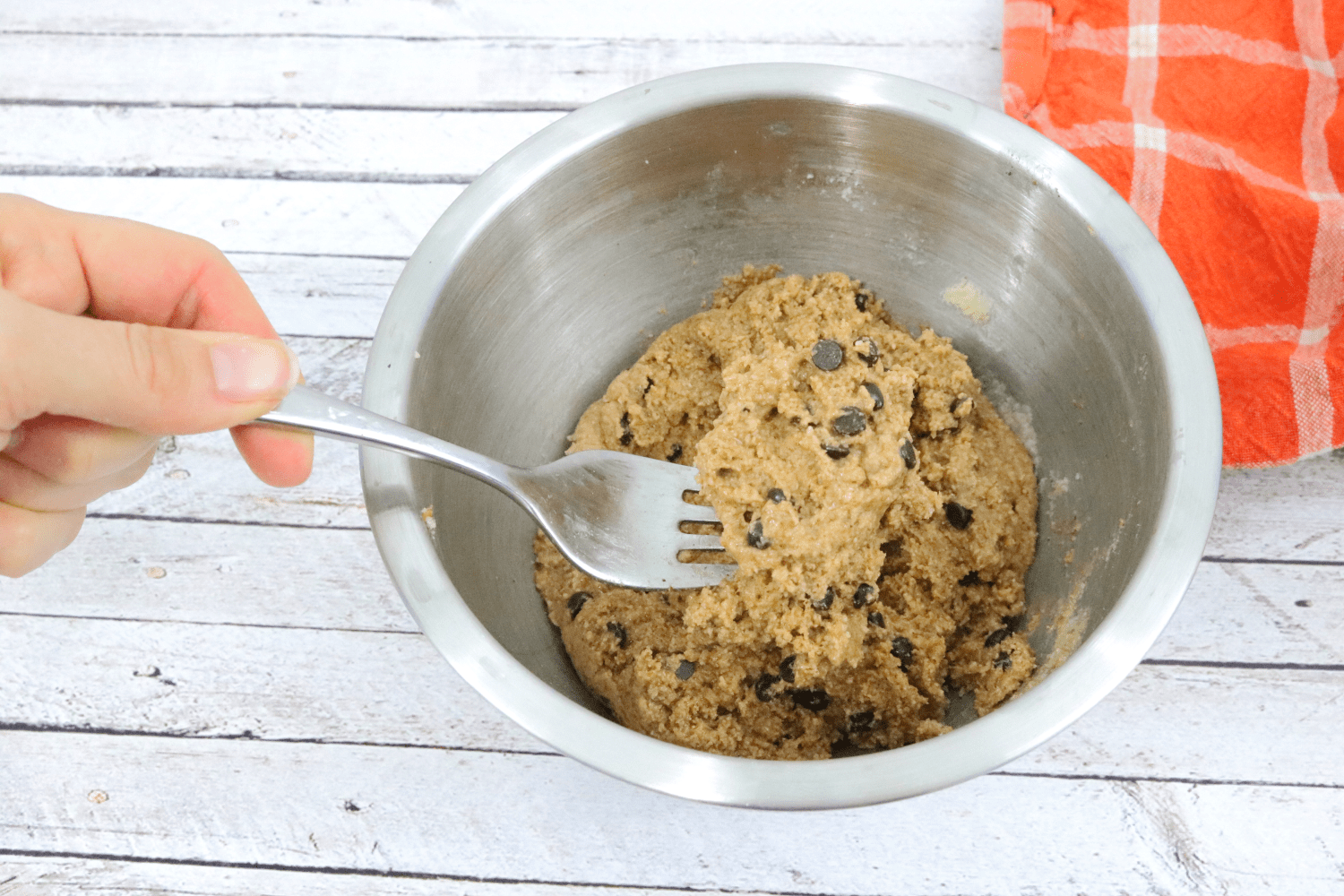 Steel bowl with raw chocolate chip cookie dough being stirred with a fork
