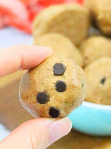 Raw cookie dough ball with chocolate chips being held between two fingers and more balls in a bowl behind them.