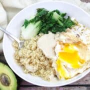 White bowl with eggs, quinoa, and bok choy with avocado beside the bowl.