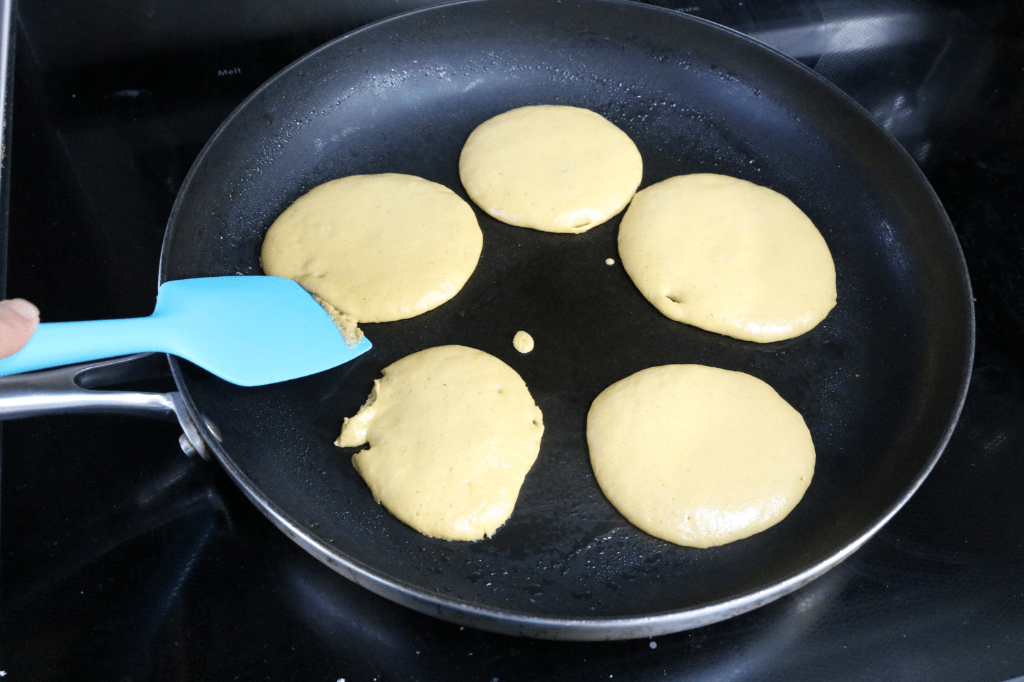 Five protein pancakes in a black pan with a blue spatula about to flip them