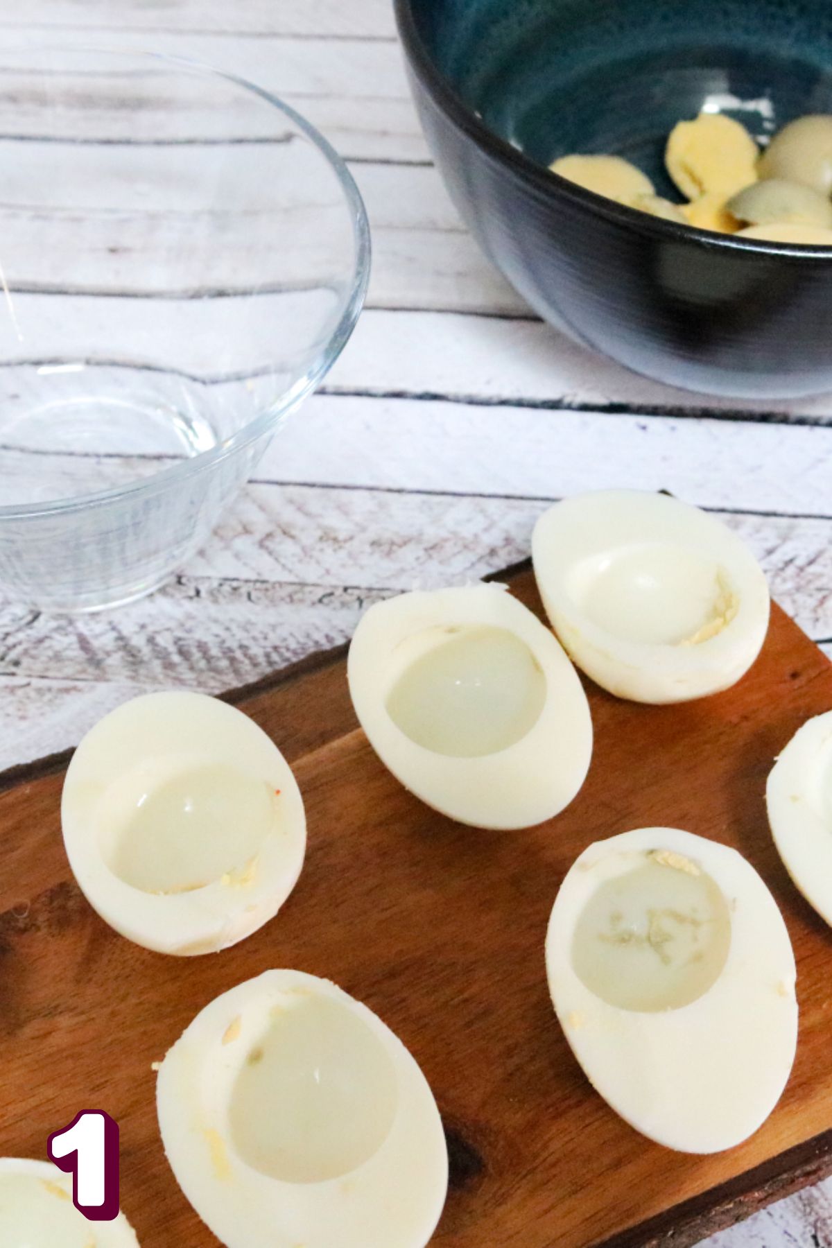 Hard boiled eggs peeled and sliced on a wooden cutting board with bowls beside it.