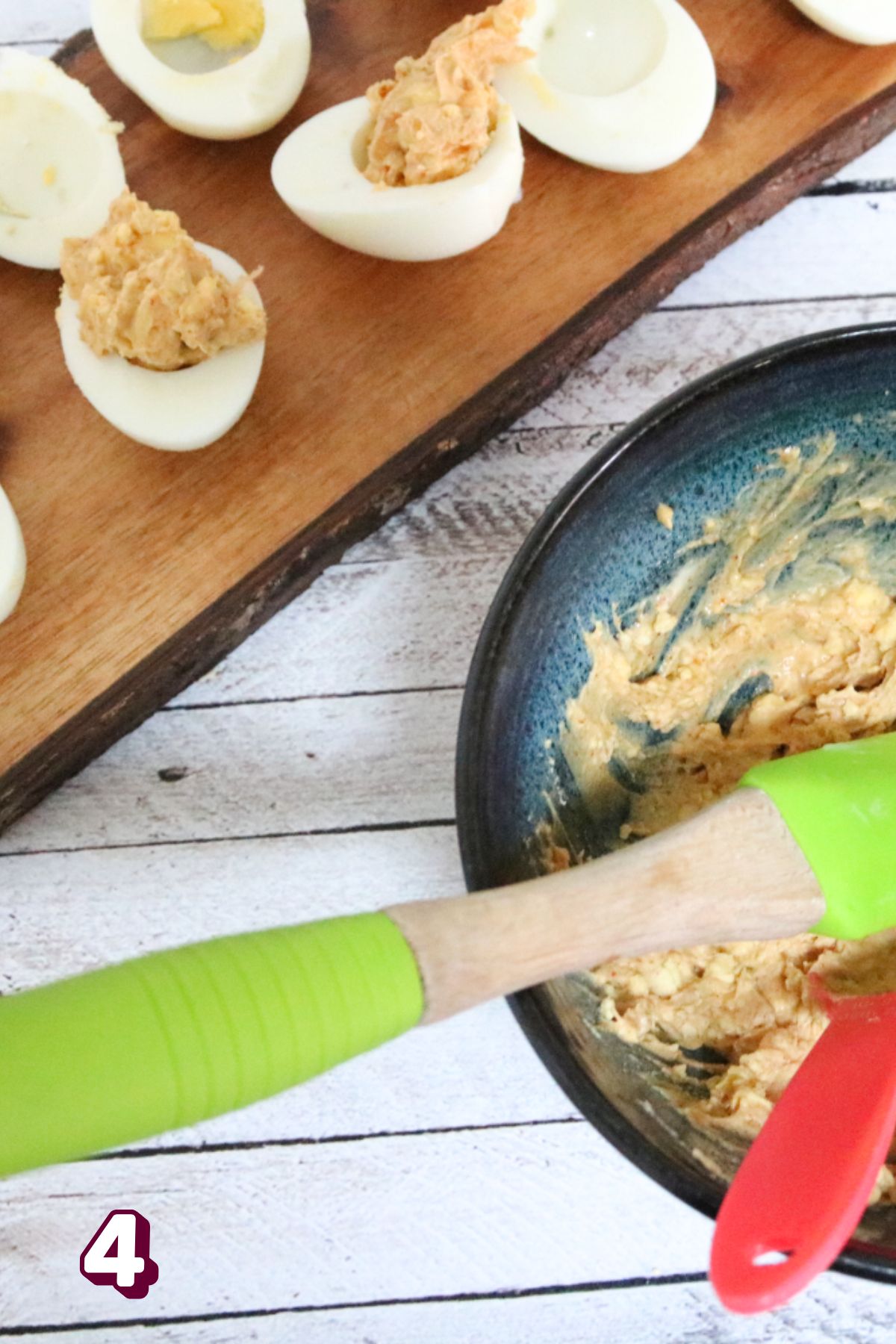 Blue bowl with deviled egg mixture in it with green spatula and hardboiled eggs behind it.