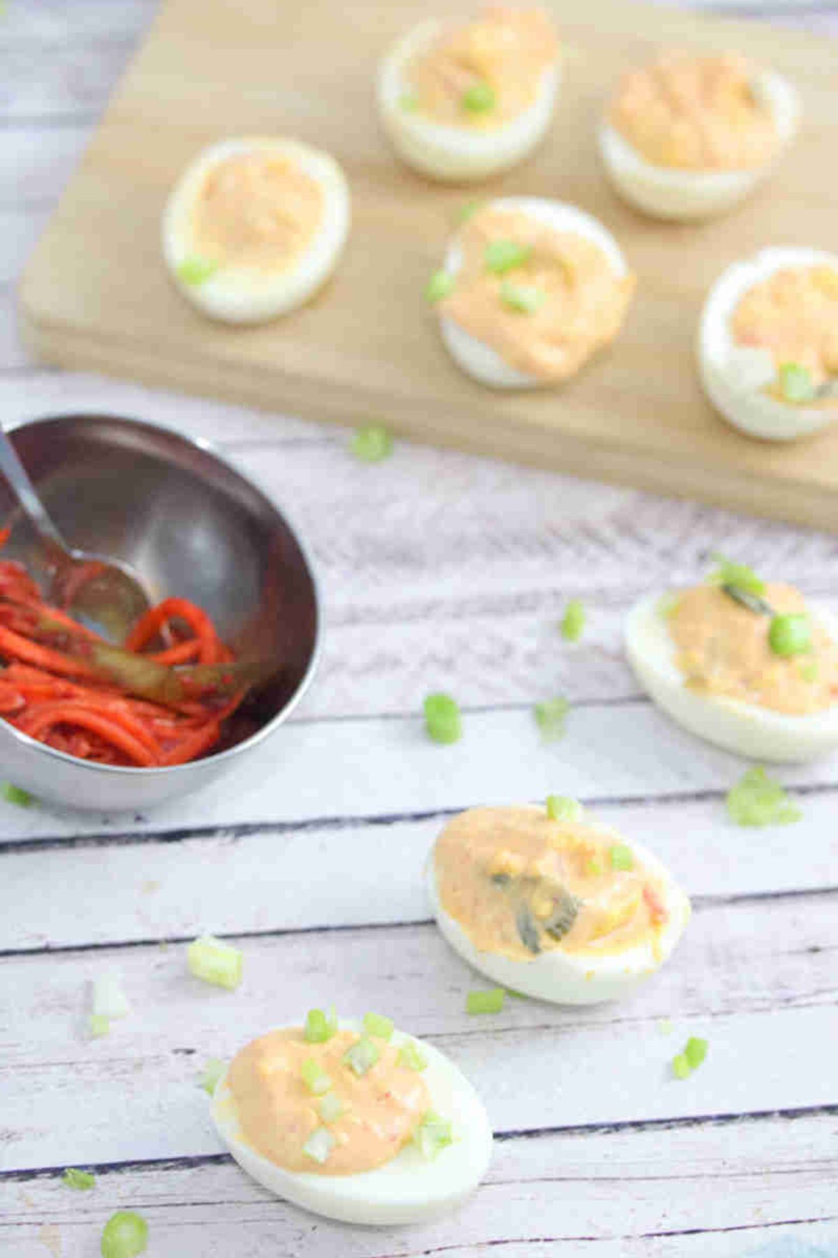 Kimchi spicy deviled eggs lined up with green onions on top and a bowl of kimchi behind them with more eggs on a cutting board.