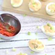 Kimchi spicy deviled eggs lined up with green onions on top and a bowl of kimchi behind them with more eggs on a cutting board.